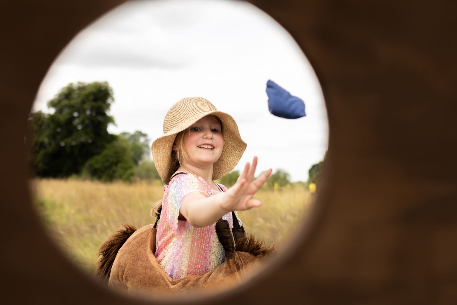 A girl with a floppy hate throwing a bean bag, picture taken through a hole framing her face